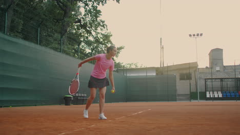 The-girl-took-her-stance-before-serving.-Smash-after-bouncing-she-hits-the-tennis-ball-near-the-ground-with-a-right-hand-A-Clay-court-with-a-green-fence.-Wide-angle-through-the-mesh.